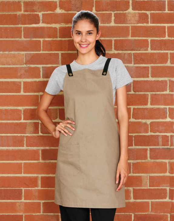A woman stands against a brick wall, wearing a beige Brunswick Bib Apron with black straps. The apron features a pocket.
