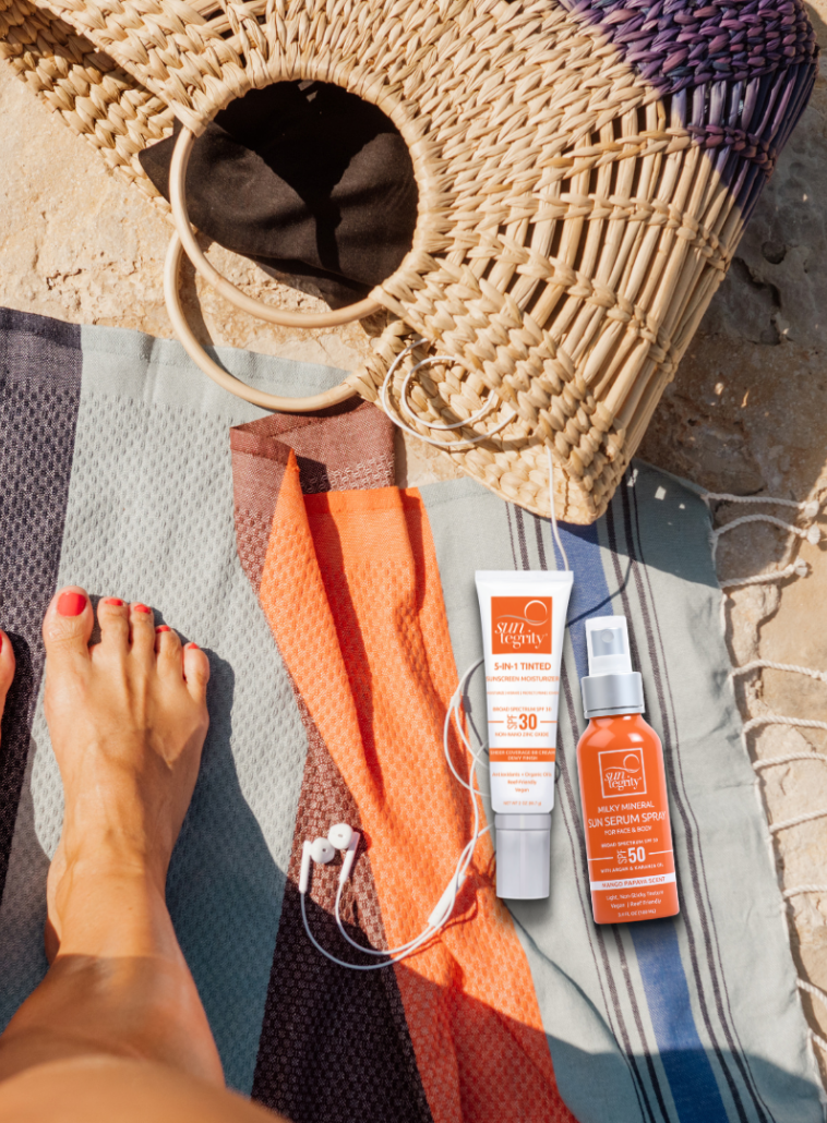 Lifestyle image of Suntegrity 5-in-1 Tinted Sunscreen Moisturizer and Suntegrity Milky Mineral Sun Serum lying on a beach throw next to a beach bag, headphones and a woman's feet.