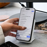 A smartphone is held in a hand, resting on a wireless charger, with a computer monitor and books in the background.