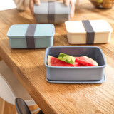A set of three rectangular lunch boxes in blue, cream, and grey, with a grey strap, containing watermelon slices.