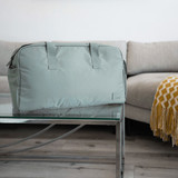 A green duffle bag sits on a glass table, with a sofa and blanket in the background. The bag has handles and a logo.
