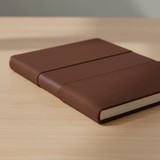 A brown notebook with a textured cover, featuring elastic bands on the front, placed on a wooden surface.
