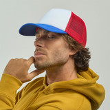 A man wearing a red and blue trucker cap, with a white front panel, poses thoughtfully in a yellow hoodie.