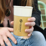 A bamboo coffee cup in natural and cream colours, held by a person with a casual outfit. Features branding on the side.