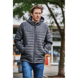 A man wearing a dark grey quilted hoody, walking outdoors surrounded by greenery. The hoody features a logo.