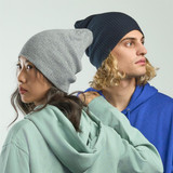 A grey and a navy beanie displayed on two models, featuring a slouchy design and a branding logo.