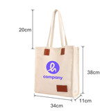 A cream canvas bag with a magnetic snap, leather accents, and a logo on the front, measuring 20cm by 34cm by 38cm.
