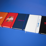 A collection of A6 notebooks in red, navy blue, white, and black, featuring various logos and designs.