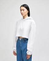 A white cropped hoodie with a drawstring hood, paired with blue jeans, featuring a logo on the sleeve.