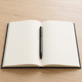 A black ballpoint pen rests on an open, blank, black-covered notebook against a wooden background.