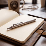 A silver 4-in-1 pen rests on an open notebook, with a coffee cup and glasses in the background.