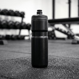 A drink bottle in black, standing upright on a gym floor with weights in the background.