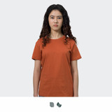 A woman stands wearing an orange TRENDSWEAR original women's T-shirt. The shirt has a simple design and a logo.