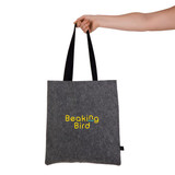 A grey felt tote bag with black straps is held by a hand, featuring a logo design.