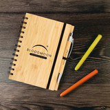 A bamboo notebook with a black elastic band, accompanied by a silver pen and colourful highlighters in orange, green, and yellow.