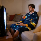 A navy blue wearable blanket with a hood, featuring colourful patterns, worn by a person sitting on a couch while watching TV.