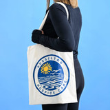 A cream Calico tote bag featuring a circular logo with sun and waves, held by a person against a blue background. A cream Calico tote bag featuring a circular logo with sun and waves, held by a person against a blue background.