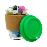 A clear glass coffee cup with a cork band and a green silicone lid, filled with colourful jelly beans.