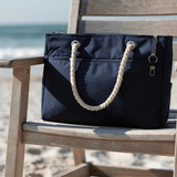 A navy waterproof beach bag with rope handles rests on a wooden chair by the shore. Features a logo.