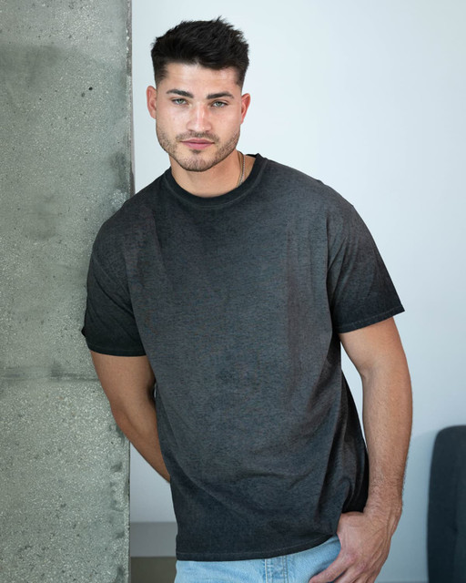 A model poses in a dark grey Colortone® oil wash t-shirt against a concrete background. The shirt features a logo.