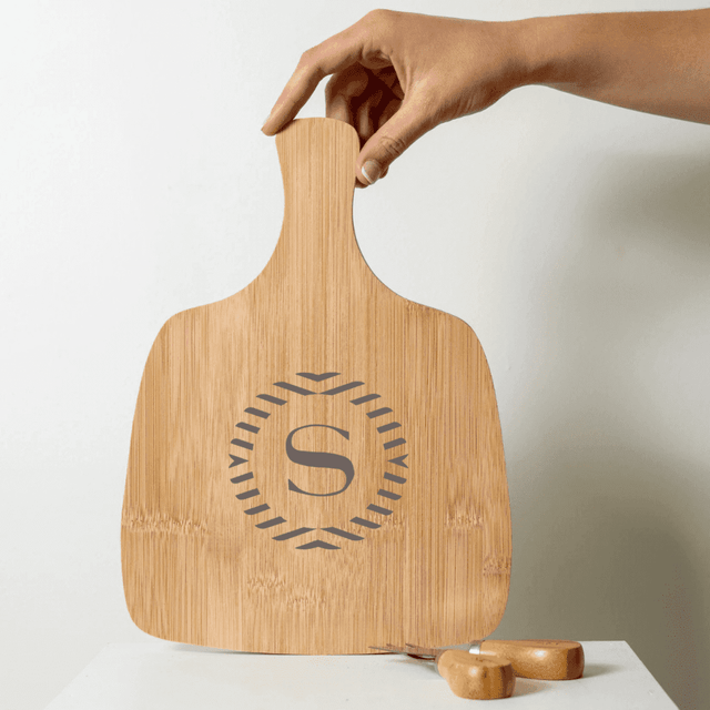A bamboo cheeseboard with an engraved logo and a hand holding it. Two wooden cheese knives are nearby.