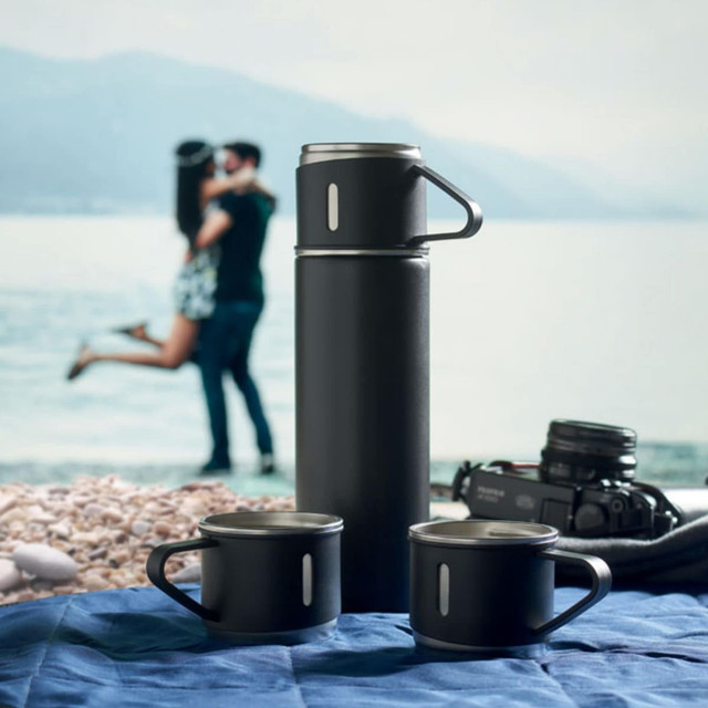 A drink bottle and two black mugs on a blue surface, with a scenic backdrop. Contains a logo.