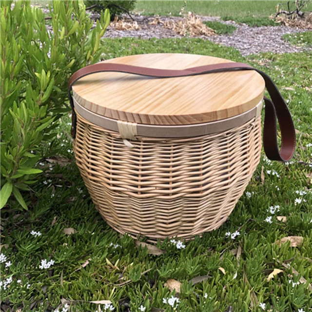 A round wicker picnic cooler basket with a wooden lid and a leather strap, set on grass with white flowers. A round wicker picnic cooler basket with a wooden lid and a leather strap, set on grass with white flowers.