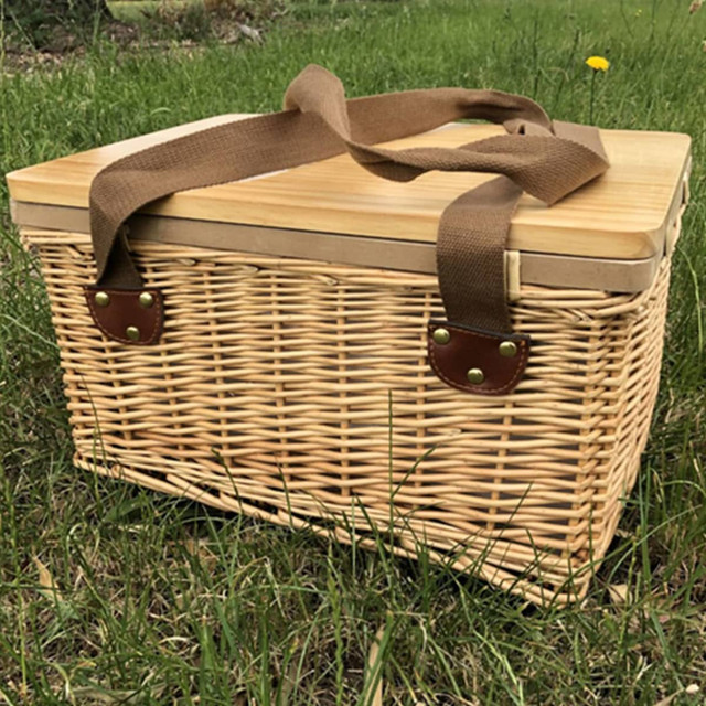 A wicker picnic cooler basket in natural tones with a bamboo lid and brown fabric handles, resting on grass. A wicker picnic cooler basket in natural tones with a bamboo lid and brown fabric handles, resting on grass.