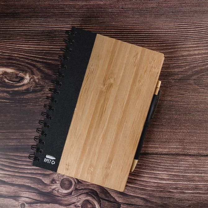 A spiral-bound A5 notebook with a bamboo and black cover, featuring a branding logo and a pen attached.