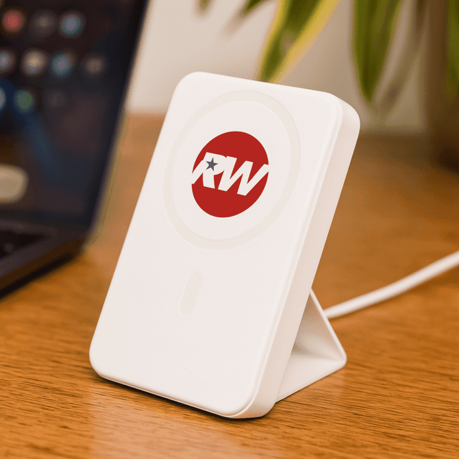A white wireless charging power bank with a logo, positioned on a wooden surface next to a laptop.