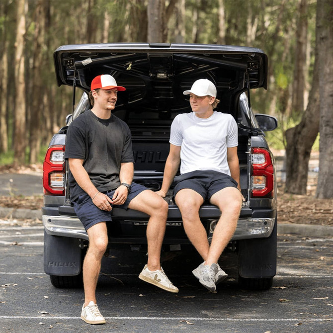 Two young men sitting on the tailgate of a truck, wearing a red and white Turbine Trucker Cap and a white cap.
