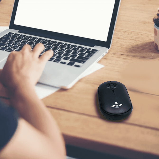 A black Slim Wireless Travel Mouse rests on a desk next to a laptop, with a hand positioned on the mouse.