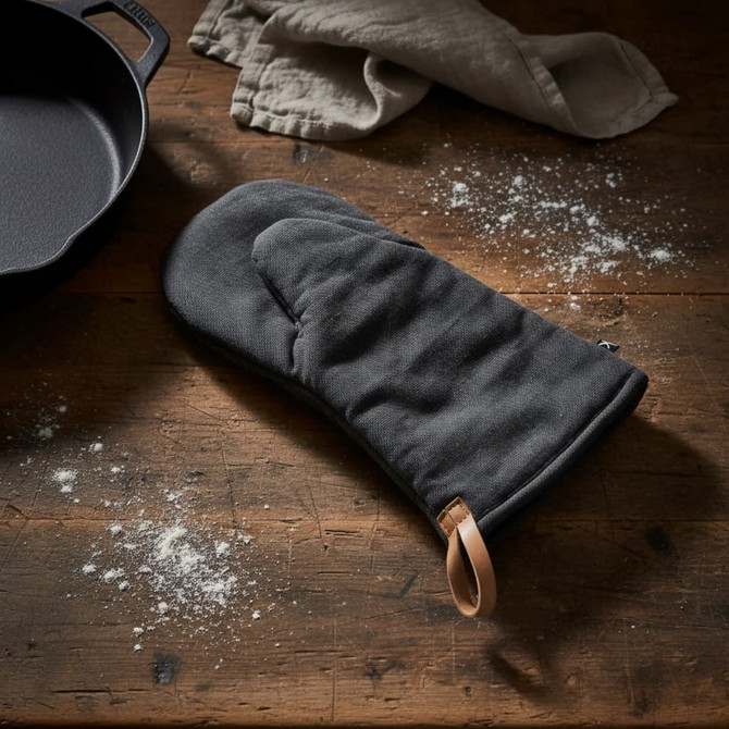 A black oven mitt with a leather loop, resting on a wooden surface sprinkled with flour next to a cast iron pan.