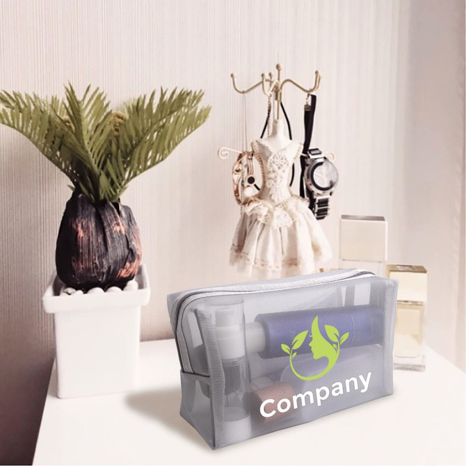 A grey nylon mesh cosmetic bag sits on a table next to a potted plant and decorative items, featuring a logo.