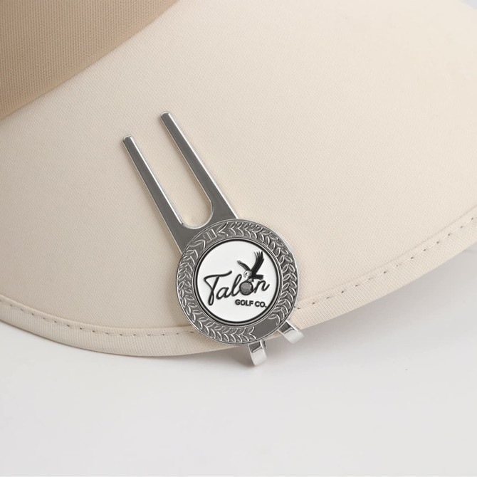 A silver golf divot tool with a ball marker attached, resting on a beige cap. It features a logo design.