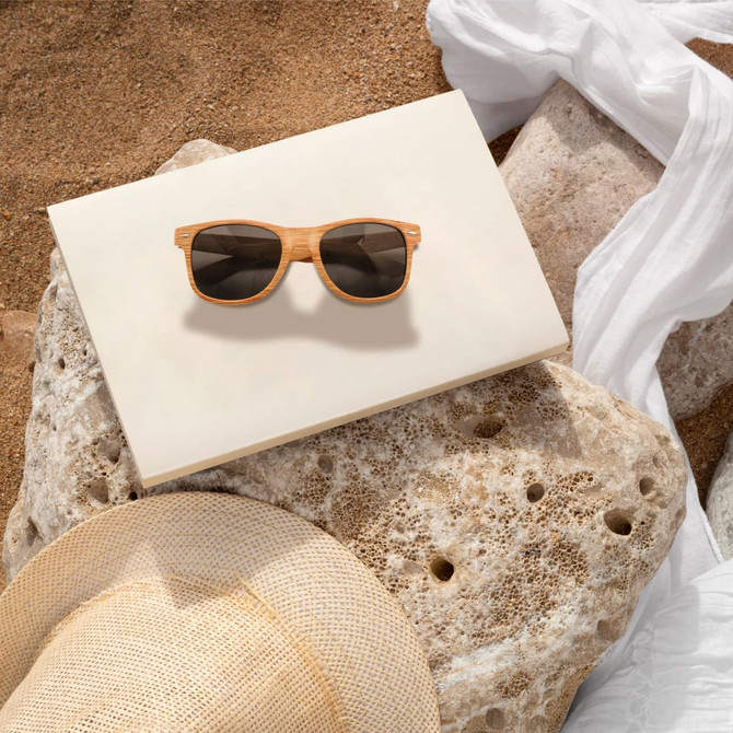 Wooden sunglasses with brown lenses resting on a light-coloured surface, surrounded by textured stones and a straw hat.