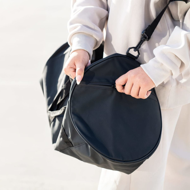 A black RPET duffle bag is held by a person, featuring a round zipped compartment and an adjustable strap.