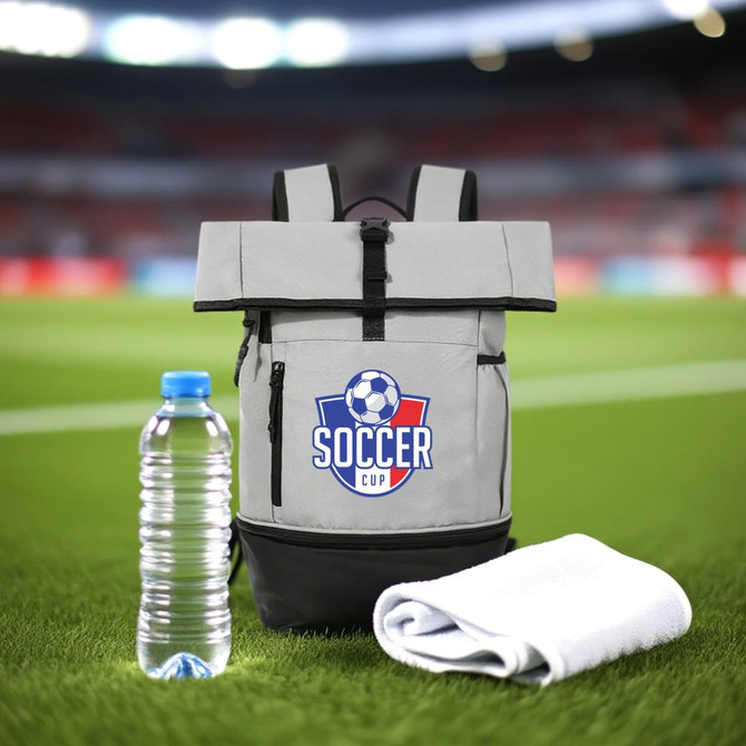 A grey and black sports roll top backpack with a logo, accompanied by a water bottle and a white towel on grass.