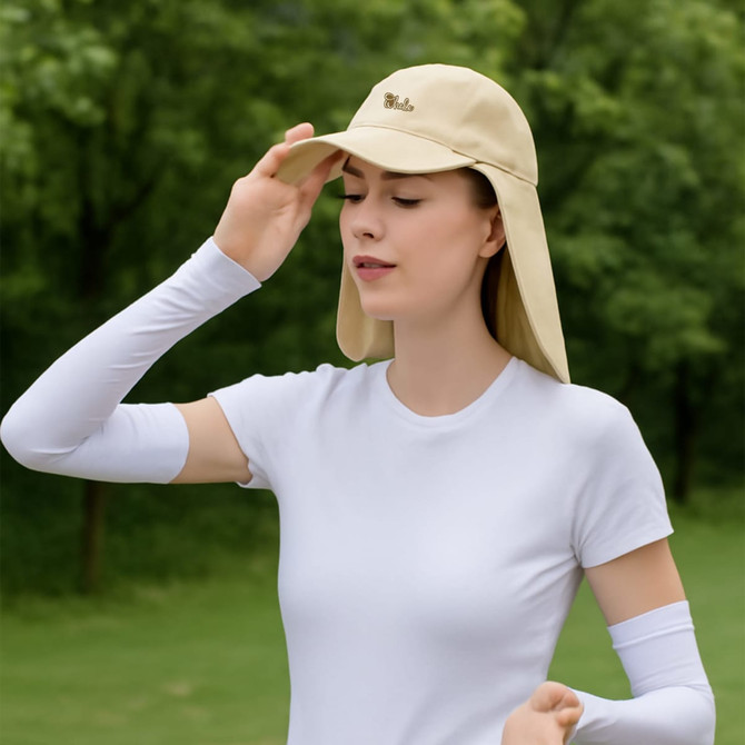 A beige baseball cap with a neck flap, worn by a woman, set against a green outdoor background. The cap has a logo.