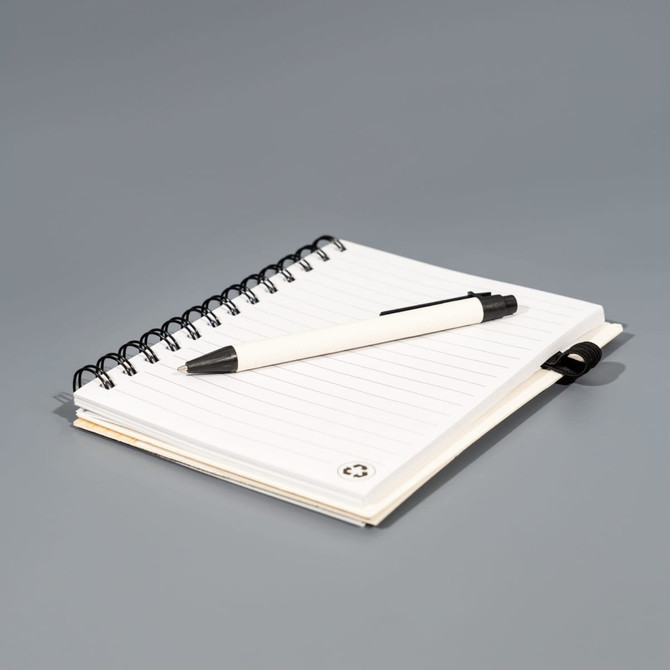 A white spiral-bound notebook with lined pages sits on a grey surface alongside a black pen. It features a logo on the cover.