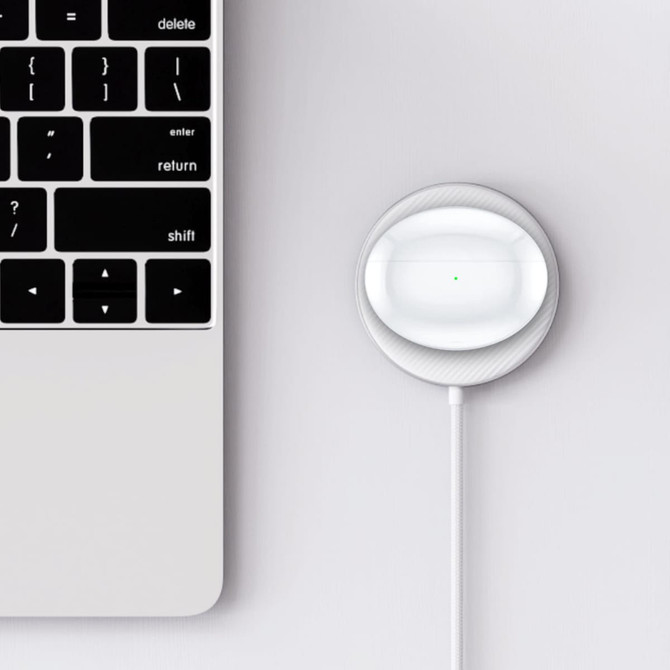 A white charging case for wireless earbuds sits next to a laptop keyboard, connected by a charging cable.