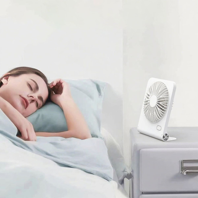 A white mini desk fan on a bedside table next to a person sleeping, with a softly lit bedroom background.