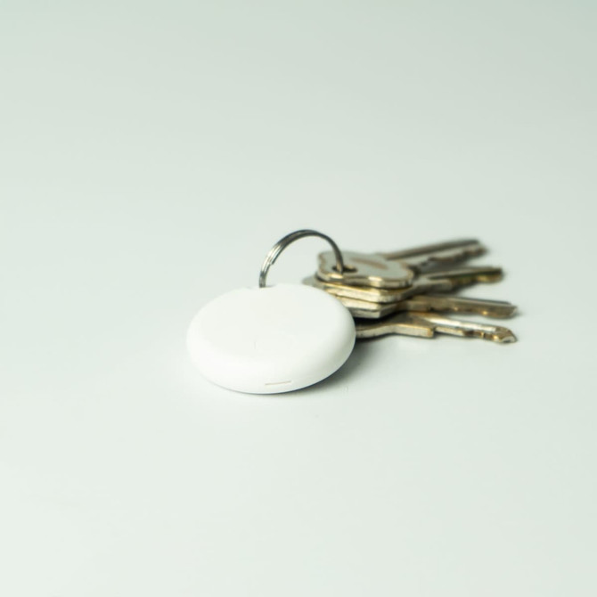 A white dual system item tracker sits alongside a set of metal keys on a light background. The tracker has a keyring attached.