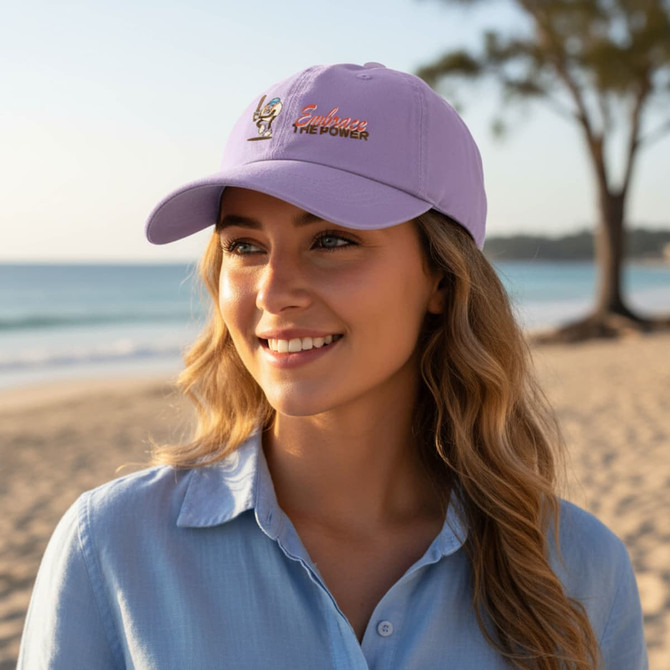 A lilac baseball cap with a logo, worn by a smiling woman on a beach, with a scenic ocean view in the background.