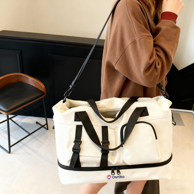 A white weekender bag with black straps, featuring a front pocket and a logo, held by a person wearing a brown sweater.