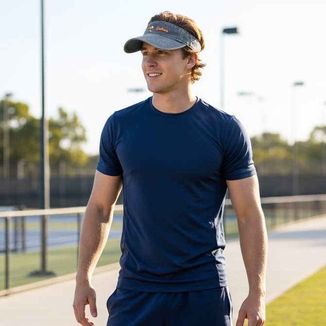 A washed sun visor in grey, worn by a man in a navy athletic shirt, set against an outdoor sports backdrop.