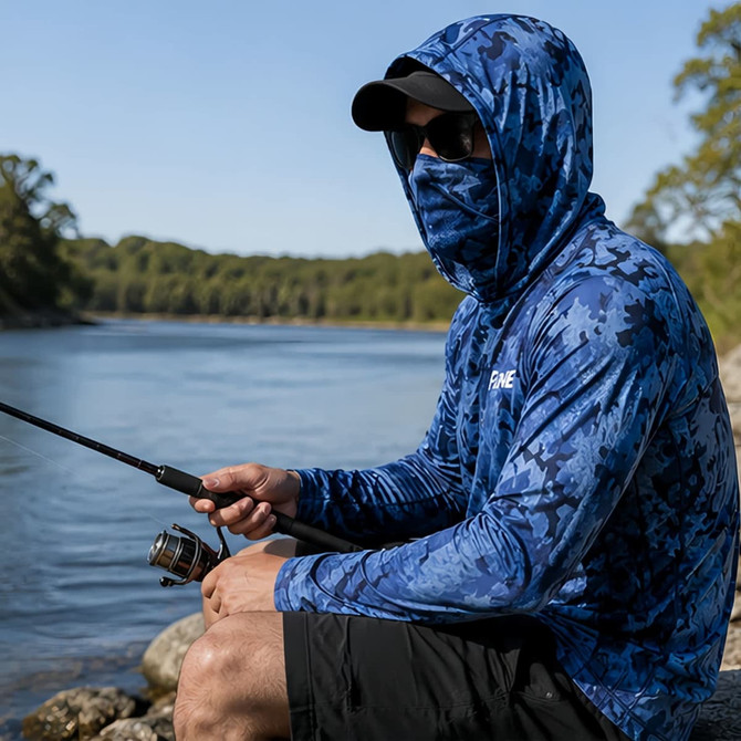 A blue camo fishing hoodie made from polyester and spandex, featuring a pocket and a logo, worn by a person by the river.