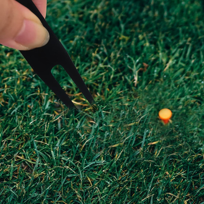 A black golf divot tool is held above a golf tee on green grass. The image features vibrant green hues.