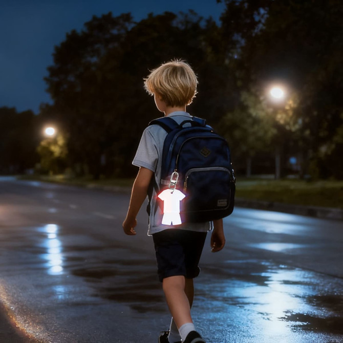 A reflective keychain in the shape of a person hangs from a child's backpack as he walks on a wet street at night.
