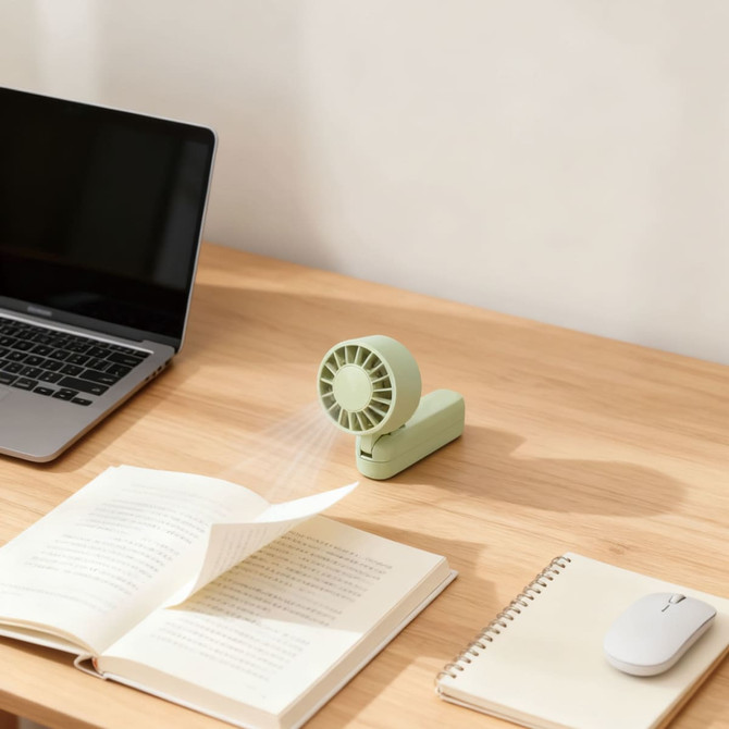 A foldable handheld fan in mint green placed on a wooden desk alongside a laptop and open books.